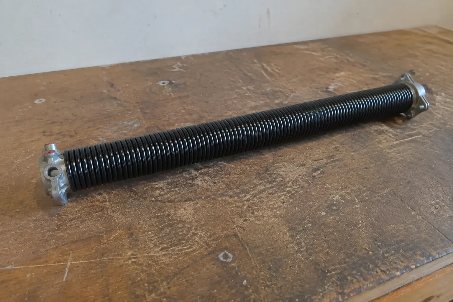 Detached black coil spring with metal end fittings on a workbench. — Torsion Spring Replacement, Whitby