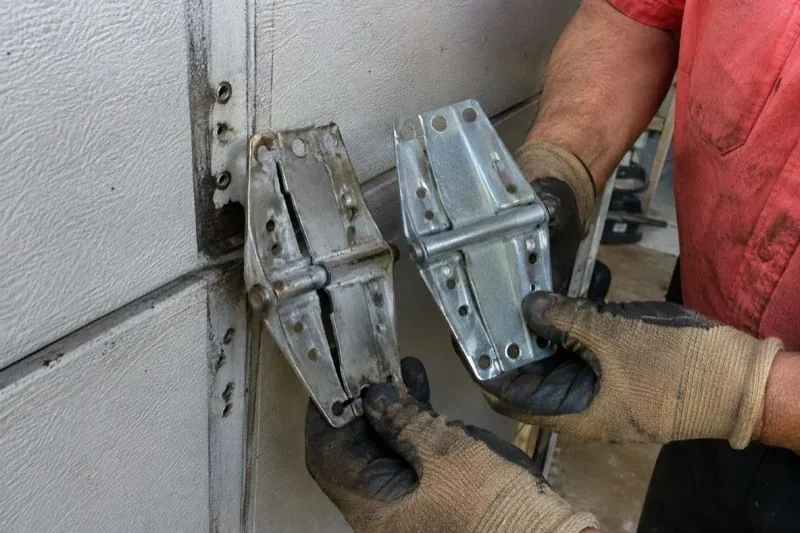 Gloved hands holding two metal garage-door hinges side by side, one discolored with a visible crack and the other bright silver. — Garage Door Hinge Replacement, Whitby