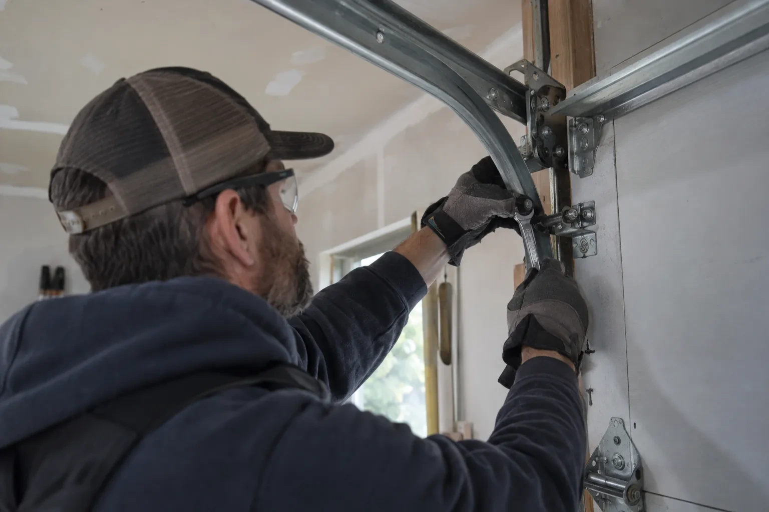 Technician in a mesh cap and safety glasses holding a wrench to a bracket at the bend of the metal door track.