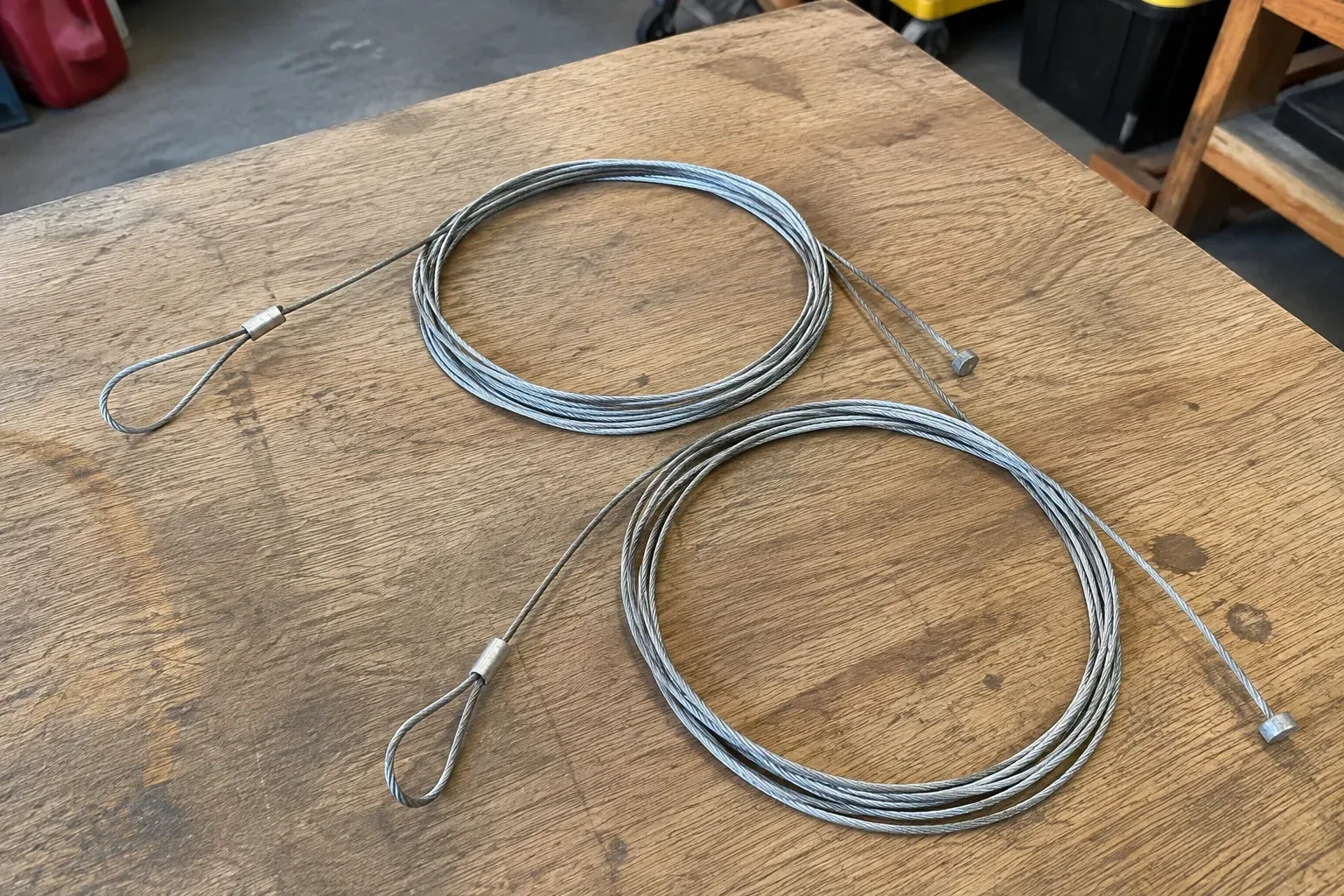 Two coiled steel cables with loop and stop ends on a workbench. — Garage Door Cable Replacement, Whitby